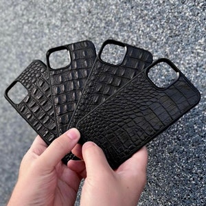 May include: A set of four black phone cases featuring a textured crocodile skin pattern. Each case has a rectangular camera cutout in the upper left corner and a slim, rigid frame design. The cases are arranged in a fan shape, displaying the pattern.