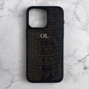 May include: A black smartphone case featuring a dark brown, textured crocodile-patterned inlay on the back. The case includes a square camera cutout and the letters OL embossed in a light, metallic-toned font centered on the upper portion.