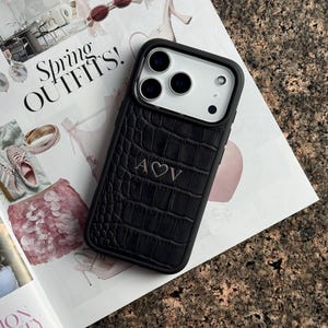 May include: A black smartphone case with a textured crocodile skin pattern and a white camera module area. The center of the case features the engraved letters A and V separated by a heart symbol. The case has a black frame and a rectangular shape.