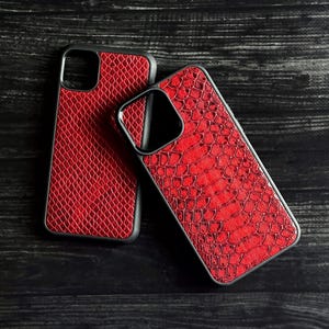 Pode incluir: Duas capas de telefone vermelhas com um padr&atilde;o texturizado de pele de cobra. As capas t&ecirc;m bordas pretas e s&atilde;o exibidas em uma superf&iacute;cie de madeira escura. As capas s&atilde;o projetadas para proteger o telefone.