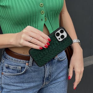 Op de afbeelding: Een rechthoekig smartphonehoesje met een donkergroen gestructureerd oppervlak met een patroon dat lijkt op reptielenschubben. Het hoesje heeft een zwarte rand en een uitsparing voor een camerasysteem met drie lenzen.