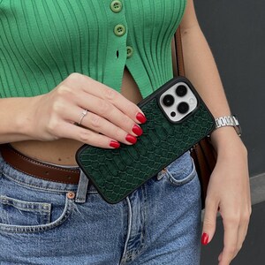 Pode incluir: Uma capa de telefone verde escuro com um padr&atilde;o texturizado semelhante &agrave; pele de r&eacute;ptil. A capa tem uma borda preta e cobre a c&acirc;mera do telefone. A pessoa na imagem est&aacute; usando um top verde e jeans azuis.