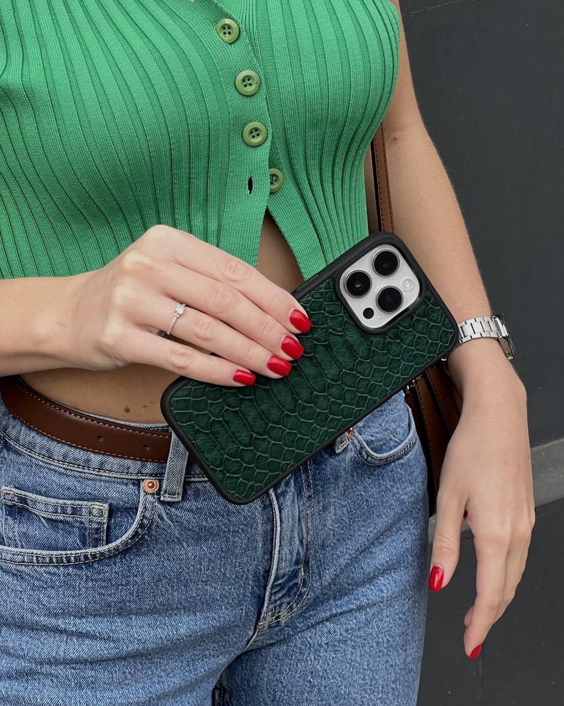 Pode incluir: Uma capa de telefone verde escuro com um padr&atilde;o texturizado semelhante &agrave; pele de r&eacute;ptil. A capa tem uma borda preta e cobre a c&acirc;mera do telefone. A pessoa na imagem est&aacute; usando um top verde e jeans azuis.