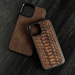 Pode incluir: Duas capas de telefone castanhas com textura de pele de cobra e bordas pretas. As capas, de tamanhos diferentes, s&atilde;o provavelmente para diferentes modelos de telefone. Est&atilde;o sobre uma superf&iacute;cie de madeira escura.