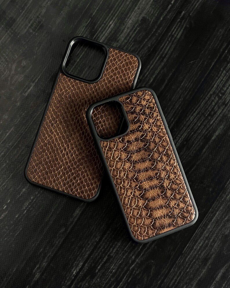 Pode incluir: Duas capas de telefone castanhas com textura de pele de cobra e bordas pretas. As capas, de tamanhos diferentes, s&atilde;o provavelmente para diferentes modelos de telefone. Est&atilde;o sobre uma superf&iacute;cie de madeira escura.