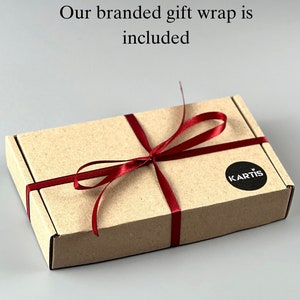Pode incluir: Uma caixa retangular de cart&atilde;o amarrada com uma fita bord&ocirc;. A caixa tem um adesivo circular preto com a palavra "KARTIS". O texto "Our branded gift wrap is included" est&aacute; acima da caixa.