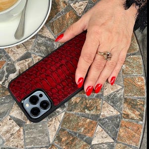Op de afbeelding: Een smartphonehoesje met een dieprood, getextureerd kunstlederen alligatorhuidpatroon op het achterpaneel. Het hoesje heeft een zwart frame en een uitsparing voor de drievoudige cameralens van een moderne smartphone.