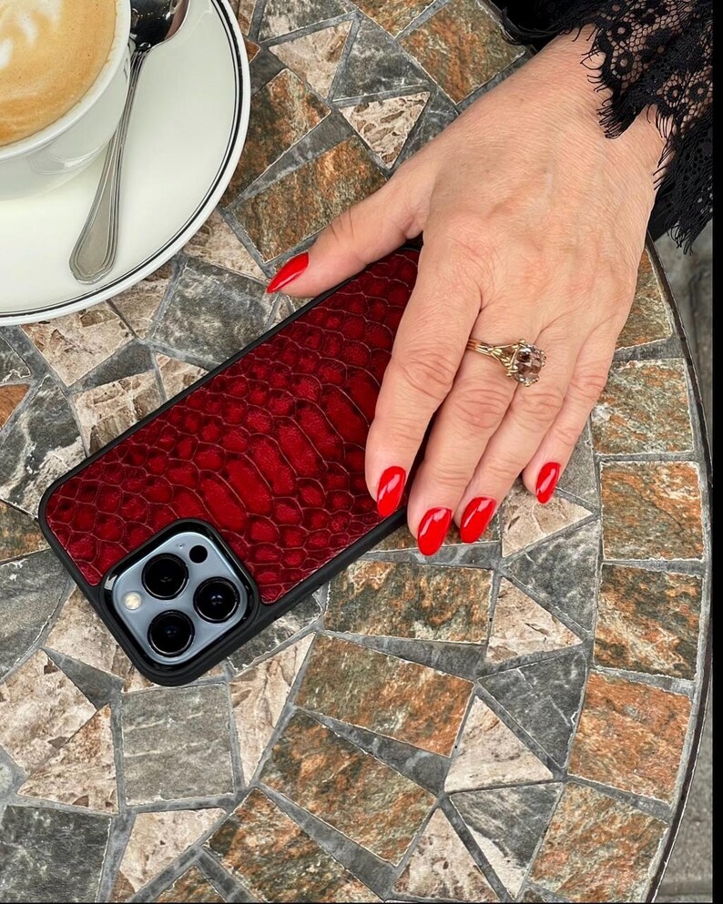Op de afbeelding: Een smartphonehoesje met een dieprood, getextureerd kunstlederen alligatorhuidpatroon op het achterpaneel. Het hoesje heeft een zwart frame en een uitsparing voor de drievoudige cameralens van een moderne smartphone.