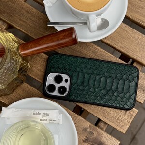 Op de afbeelding: Een rechthoekig smartphonehoesje met een donkergroen, getextureerd achterpaneel met slangenleerpatroon, een zwart frame en een ronde uitsparing voor de camera, geplaatst op een houten oppervlak naast een witte koffiekop en een glas.