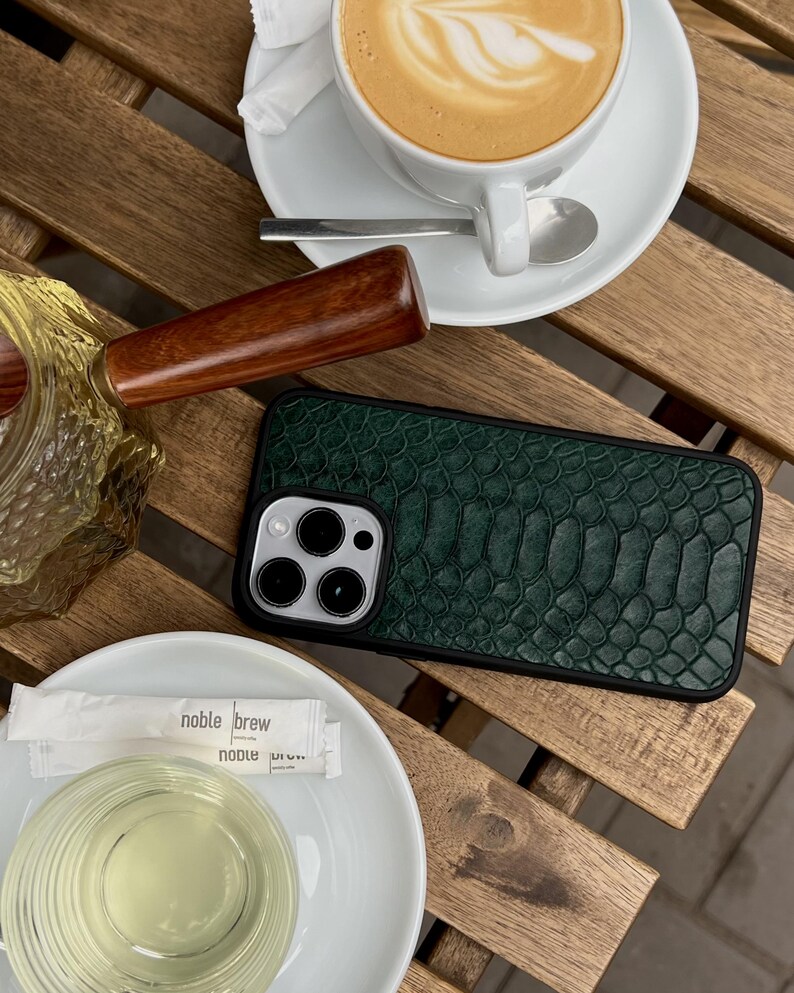 Op de afbeelding: Een rechthoekig smartphonehoesje met een donkergroen, getextureerd achterpaneel met slangenleerpatroon, een zwart frame en een ronde uitsparing voor de camera, geplaatst op een houten oppervlak naast een witte koffiekop en een glas.