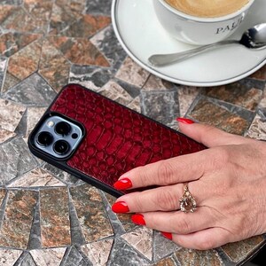 Op de afbeelding: Een smartphonehoesje met een dieprood, getextureerd achterpaneel met slangenleerpatroon, een zwart frame en een uitsparing voor de drievoudige cameralens. Het hoesje heeft een strak, modern ontwerp dat het apparaat beschermt.