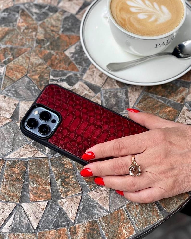 Op de afbeelding: Een smartphonehoesje met een dieprood, getextureerd achterpaneel met slangenleerpatroon, een zwart frame en een uitsparing voor de drievoudige cameralens. Het hoesje heeft een strak, modern ontwerp dat het apparaat beschermt.