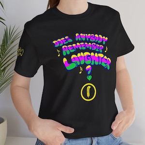 May include: A black short-sleeve t-shirt featuring the text DOES ANYBODY REMEMBER LAUGHTER in a wavy, multi-colored font with musical notes, a green heart, and a yellow circle containing a feather icon printed on the front chest area.