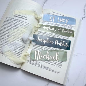May include: Four colorful bookmarks with different saint names printed in white. The bookmarks are made of clear acrylic with a painted background. The names are St. Lucy, Anthony of Padua, Josephine Bakhita, and Michael.