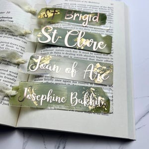 May include: Set of four green and gold resin bookmarks with the names Brigid, St. Clare, Joan of Arc, and Josephine Bakhita written in gold lettering.