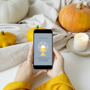 May include: A smartphone displays a religious graphic with the text "Praying for you." The graphic features a gold chalice and host with radiating light rays. The phone is held in front of a white surface with pumpkins and a lit candle.