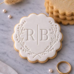 May include: A round scalloped sugar biscuit topped with smooth white icing featuring an embossed design of the letters R and B separated by a vertical line, framed by a delicate botanical wreath pattern. A stack of plain biscuits is visible nearby.