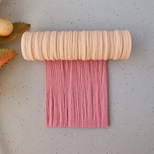 May include: A light peach-coloured, ridged cylinder sits atop a textured, rectangular piece of pink material. The pink material has vertical lines. The background is a light grey colour.