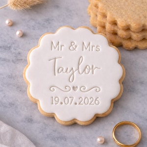 Peut inclure: Un biscuit décoré avec du glaçage blanc, portant l'inscription "Mr & Mrs Taylor" et la date "19.07.2026". Le biscuit a un bord festonné. D'autres biscuits et une alliance dorée sont visibles.
