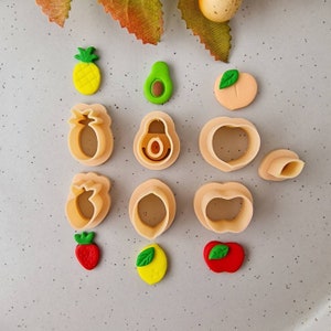 Könnte beinhalten: Eine Sammlung von Ausstechern in Obstform und passenden Obstdekorationen. Die Ausstecher sind beige, und die Dekorationen umfassen eine gelbe Ananas, eine grüne Avocado, einen Pfirsich, eine rote Erdbeere, eine gelbe Zitrone und einen roten Apfel.