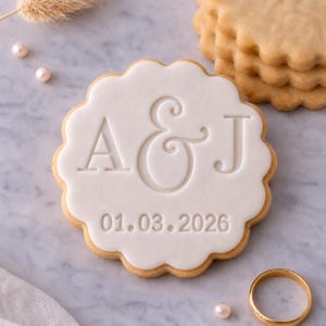 May include: A round biscuit decorated with white icing, displaying the initials "A & J" and the date "01.03.2026". The biscuit has a scalloped edge and is next to a stack of similar biscuits and a gold wedding ring.