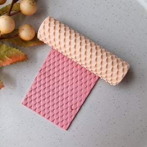 Puede incluir: Un rodillo de color melocotón claro con un patrón de escamas descansa sobre una superficie rectangular rosa con un patrón a juego. El rodillo está diseñado para crear superficies texturizadas en masa o arcilla. Vista superior.