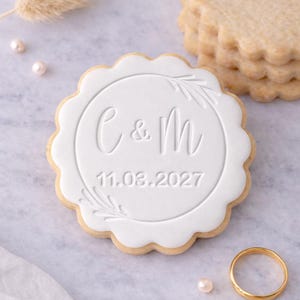 Peut inclure: Un biscuit rond glacé blanc avec des bords festonnés, gaufré des lettres "C & M" et la date "11.08.2027". Une alliance dorée et des perles sont à côté du biscuit. Une pile de biscuits similaires est en arrière-plan.