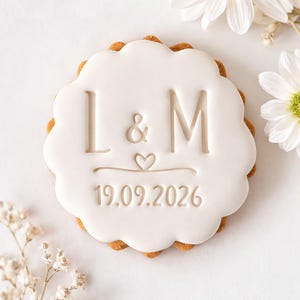 May include: A biscuit decorated with white icing, featuring the initials "L & M", an ampersand, a heart, and the date "19.09.2026". The biscuit has a scalloped edge. White flowers are arranged around the biscuit.