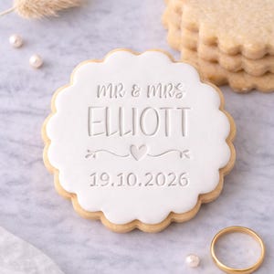 Pode incluir: Um biscoito decorado para casamento com glacê branco. Está inscrito com "MR & MRS ELLIOTT" e a data "19.10.2026". Uma aliança de ouro e outros biscoitos são visíveis. O biscoito tem uma borda ondulada e mede cerca de 8 cm de diâmetro.