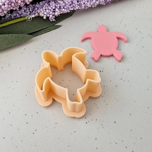 Op de afbeelding: Een licht perzikkleurige schildpadvormige koekjesuitsteker naast een roze schildpadvormige koekje. De koekjesuitsteker is gemaakt van plastic en heeft een gedetailleerde omtrek. De roze koek is gemaakt van een zacht materiaal. De achtergrond bevat paarse bloemen.