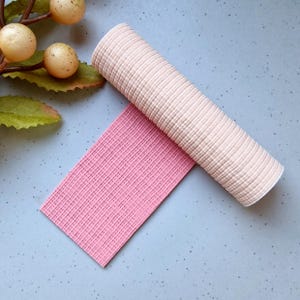 May include: A rolled beige fabric and a flat pink fabric are displayed on a light blue surface. The fabrics have a textured, waffle-like pattern. Decorative sprigs with beige orbs and green leaves are in the upper left corner.