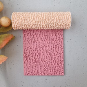 May include: A beige textured rolling pin is shown rolling over a pink surface, creating a pattern. The rolling pin has a repeating geometric design. The pink surface is rectangular. The image is shot from above.