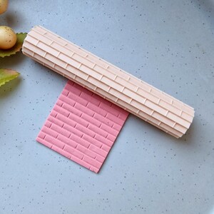 Könnte beinhalten: Ein beiger Nudelholz mit Ziegelmuster liegt über einem rosa rechteckigen Materialstück mit passendem Ziegelmuster. Das Nudelholz ist zylindrisch und hat eine strukturierte Oberfläche. Das rosa Material ist einfarbig.
