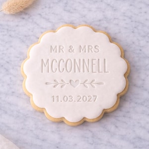Pode incluir: Um biscoito redondo com uma parte superior branca e ondulada. O biscoito está inscrito com "MR & MRS MCCONNELL" acima de um design decorativo de coração e folhas, e a data "11.03.2027". O biscoito tem uma borda marrom dourada.