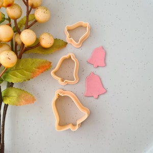 May include: Bell-shaped cookie cutters in light orange and pink. The cutters are arranged on a light grey surface, with a sprig of yellow berries and green leaves in the background. The cutters are for baking and crafting.