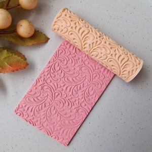 May include: A beige rolling pin with a floral pattern lies on a pink rectangular surface with the same embossed design. The rolling pin is cylindrical and has a detailed, raised floral motif. The pink surface is a rectangle with the same floral pattern.