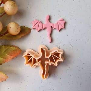May include: A pink dragon-shaped cookie cutter and a pink dragon-shaped fondant decoration are displayed on a light blue surface. The cookie cutter is a light tan color. The dragon has outstretched wings and a long tail.