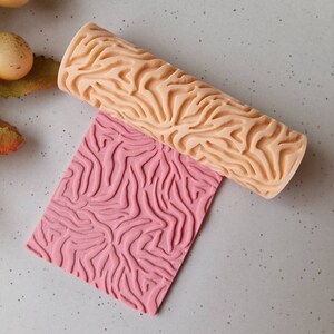 Op de afbeelding: Een licht perzikkleurige deegroller met een getextureerd zebra streepjespatroon rust boven een roze rechthoekig vel met hetzelfde patroon. De deegroller is cilindrisch en het vel is plat. De achtergrond is een lichtgrijze ondergrond.