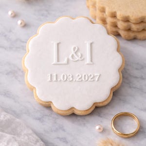 May include: A scalloped-edge sugar cookie with white icing, displaying the initials "L & I" and the date "11.03.2027". A gold wedding band and pearls are nearby. A stack of similar cookies is visible in the background.