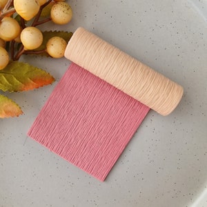 Puede incluir: Una hoja de papel crepé rosa con una superficie texturizada está parcialmente enrollada, revelando un cilindro beige texturizado. El papel y el cilindro están sobre una superficie gris claro. La imagen es un primer plano.