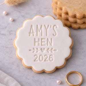 Op de afbeelding: Een rond koekje met een geschulpte rand en een laagje glad wit glazuur. Het glazuur is voorzien van de tekst AMY'S HEN, een klein hartje geflankeerd door bladtakjes en het jaartal 2026. Het koekje heeft een licht goudbruine onderkant.