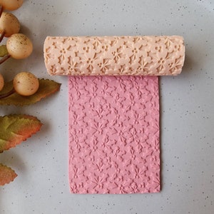May include: A beige rolling pin with a floral pattern is shown above a sheet of pink material with the same embossed design. The rolling pin is cylindrical and has a repeating pattern of flowers and berries. The pink material is rectangular.