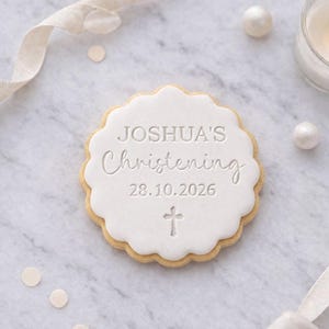 Op de afbeelding: Een ronde koek met een geschulpte rand, bedekt met witte glazuur. De glazuur is voorzien van de tekst "JOSHUA'S Christening", de datum "28.10.2026" en een kruis. De koek ligt op een marmeren oppervlak, met decoratieve elementen.