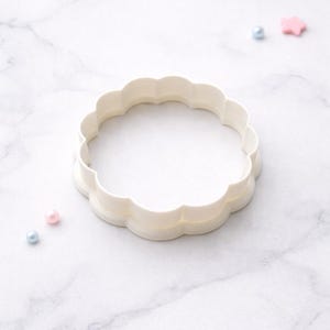 May include: A white, scalloped-edge cookie cutter in a circular shape. The cutter is made of plastic and sits on a white marble surface. Small, decorative beads in pink and blue are scattered around the cutter.