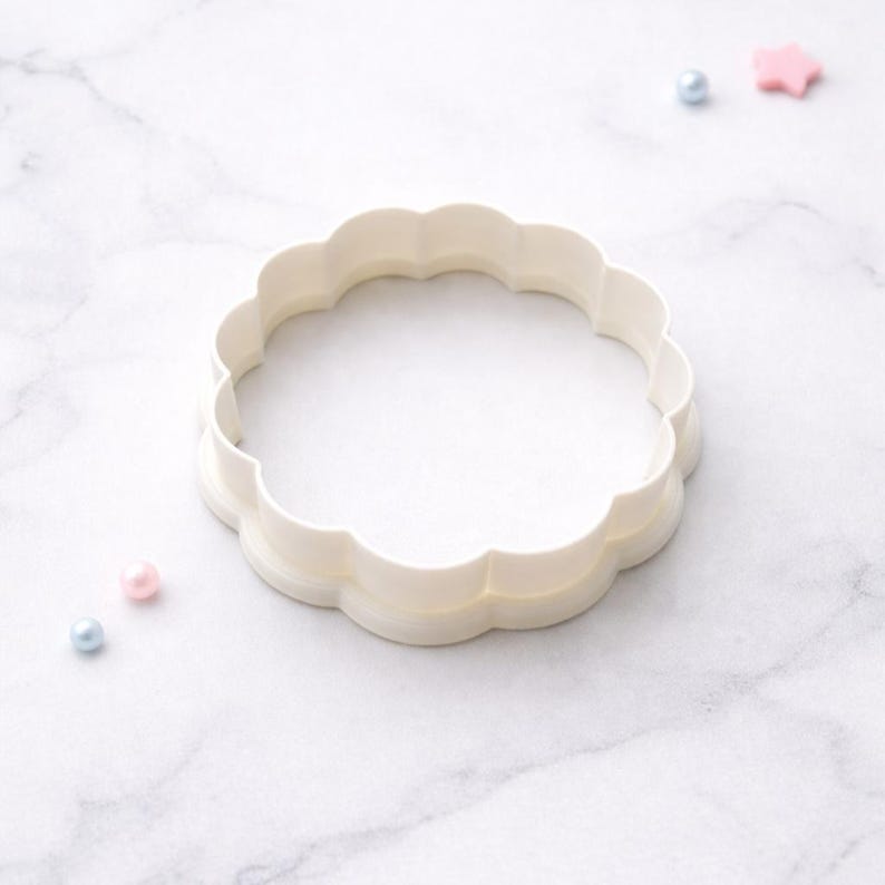 May include: A white, scalloped-edge cookie cutter in a circular shape. The cutter is made of plastic and sits on a white marble surface. Small, decorative beads in pink and blue are scattered around the cutter.
