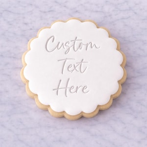 Könnte beinhalten: Ein runder Keks mit gewelltem Rand. Der Keks hat eine weiße Glasur mit den Worten "Custom Text Here" in kursiver Schrift. Der Keks liegt auf einer weißen und grauen Marmoroberfläche.