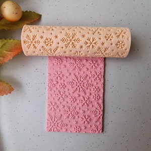 May include: A beige rolling pin with a snowflake pattern is shown rolling over a pink surface, also imprinted with snowflakes. The rolling pin is made of wood and is used for baking. The snowflakes are of varying sizes.