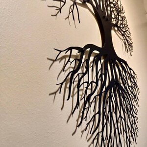 May include: Black metal tree wall art. The artwork depicts a tree with roots and branches, mounted on a light-colored wall. The tree's design casts shadows, adding depth to the wall decor. This piece is suitable for home or office decoration.