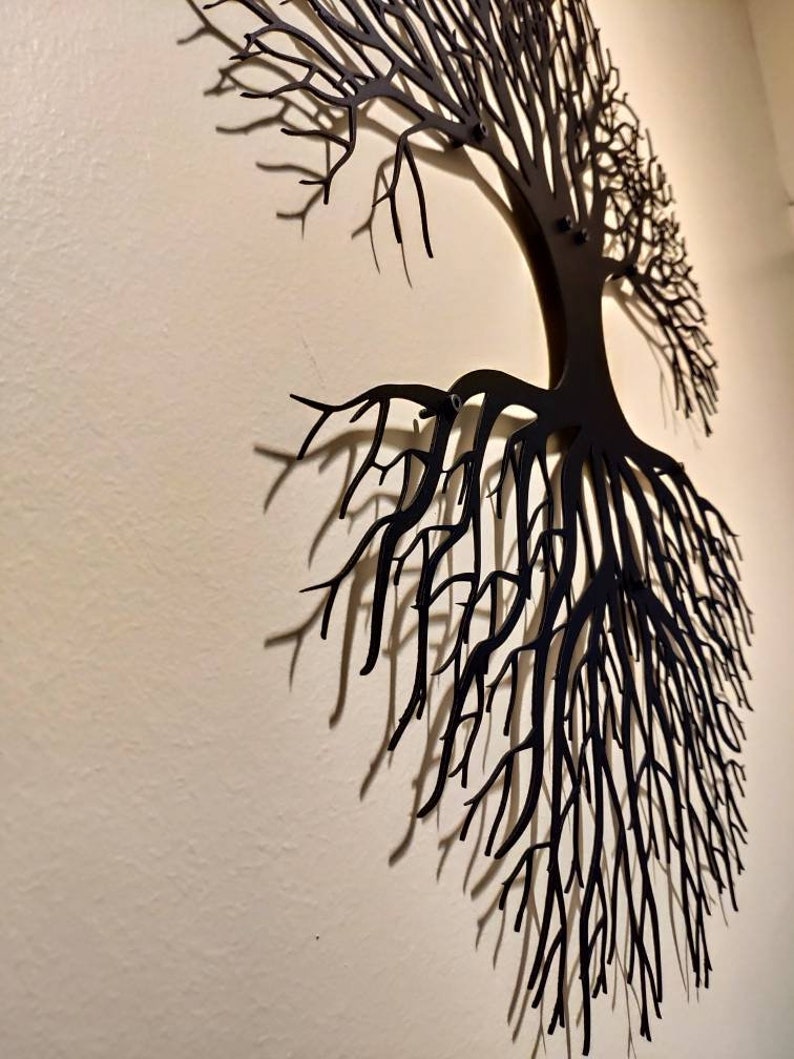 May include: Black metal tree wall art. The artwork depicts a tree with roots and branches, mounted on a light-colored wall. The tree's design casts shadows, adding depth to the wall decor. This piece is suitable for home or office decoration.