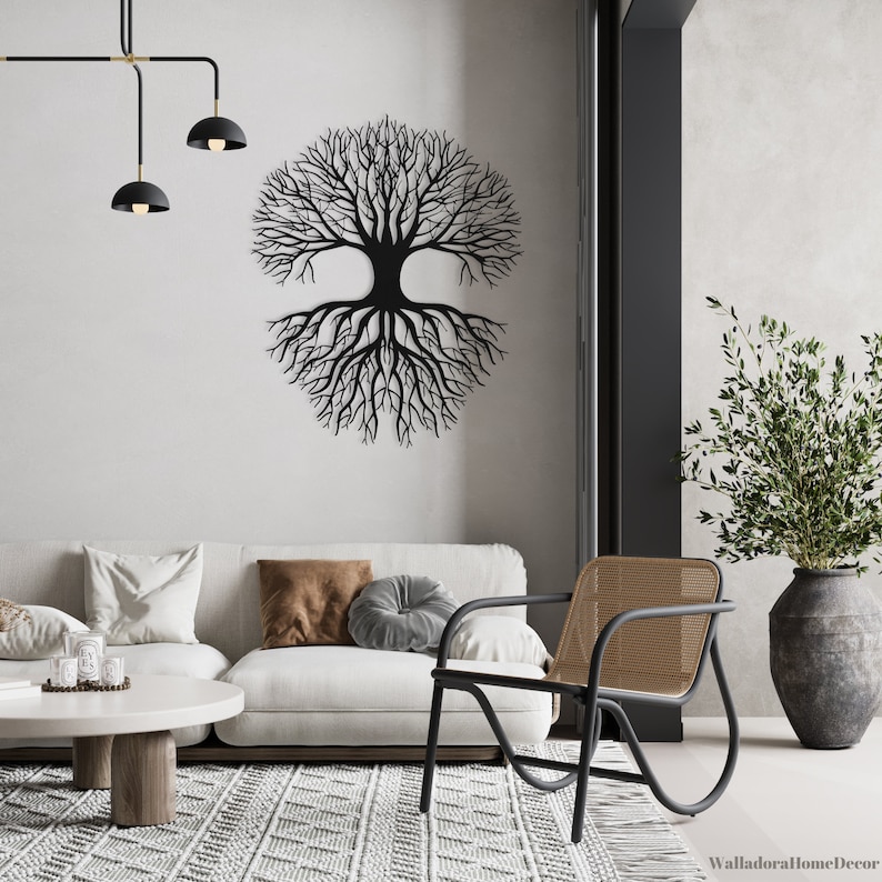 May include: A black metal tree wall decor with a large, circular, tree trunk and branches with roots extending out from the trunk. The decor is mounted on a white wall in a living room setting with a white couch, a brown throw pillow, a gray throw pillow, a black chair with a woven seat, a small round coffee table, and a green plant in a gray pot.